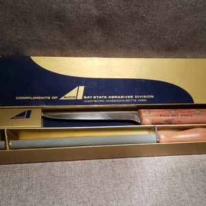 May include: A wooden-handled knife with a gray sharpening steel in a gold and blue box. The box has the text "Compliments of AVCO Bay State Abrasives Division Westboro, Massachusetts 01581" and "AVCO Bay State" on it.