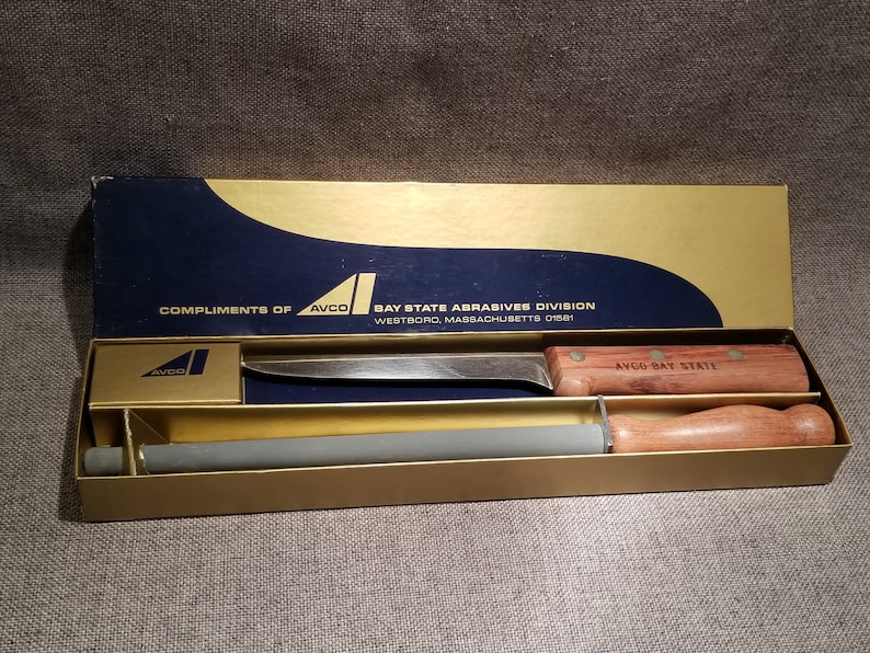 May include: A wooden-handled knife with a gray sharpening steel in a gold and blue box. The box has the text "Compliments of AVCO Bay State Abrasives Division Westboro, Massachusetts 01581" and "AVCO Bay State" on it.