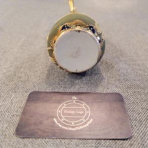 May include: A green and gold decorative bottle stopper with a white porcelain base. The stopper features a floral design with blue accents.