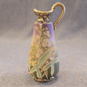 May include: A small, hand-painted porcelain pitcher with a gold handle and floral design. The pitcher is decorated with white flowers, green leaves, and gold accents on a purple and green background.