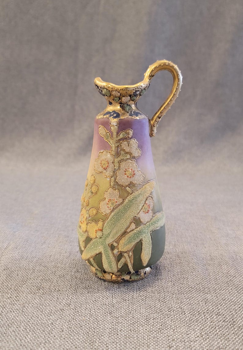 May include: A small, hand-painted porcelain pitcher with a gold handle and floral design. The pitcher is decorated with white flowers, green leaves, and gold accents on a purple and green background.