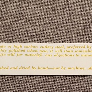 May include: A white label with gold text explaining the blade and handle of a knife. The blade is made of high carbon cutlery steel and the handle is made of imported Brazilian Rosewood. The label also suggests washing the knife by hand, not by machine. The label has the logo "AWCO" and "BAY STATE ABRASIVES DIVISION WESTBORO, MASSACHUSETTS 01581".