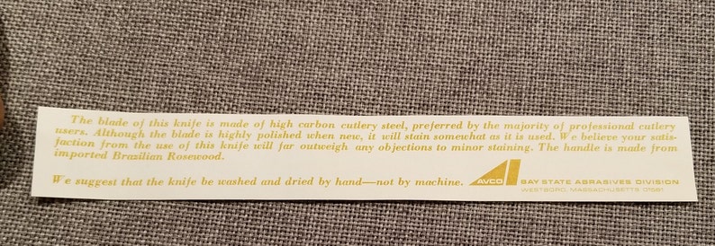 May include: A white label with gold text explaining the blade and handle of a knife. The blade is made of high carbon cutlery steel and the handle is made of imported Brazilian Rosewood. The label also suggests washing the knife by hand, not by machine. The label has the logo "AWCO" and "BAY STATE ABRASIVES DIVISION WESTBORO, MASSACHUSETTS 01581".