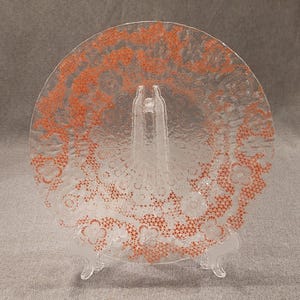 May include: A clear glass plate with an intricate orange floral pattern around the edge. The plate is displayed on a clear plastic stand, showcasing its decorative design. The plate is round and has a textured surface.