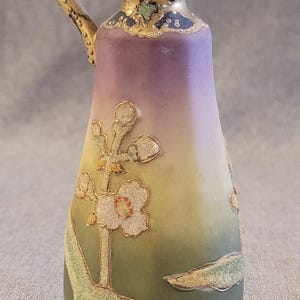 May include: A small, hand-painted porcelain pitcher with a gold handle and floral design. The pitcher has a gradient of purple, yellow, and green.