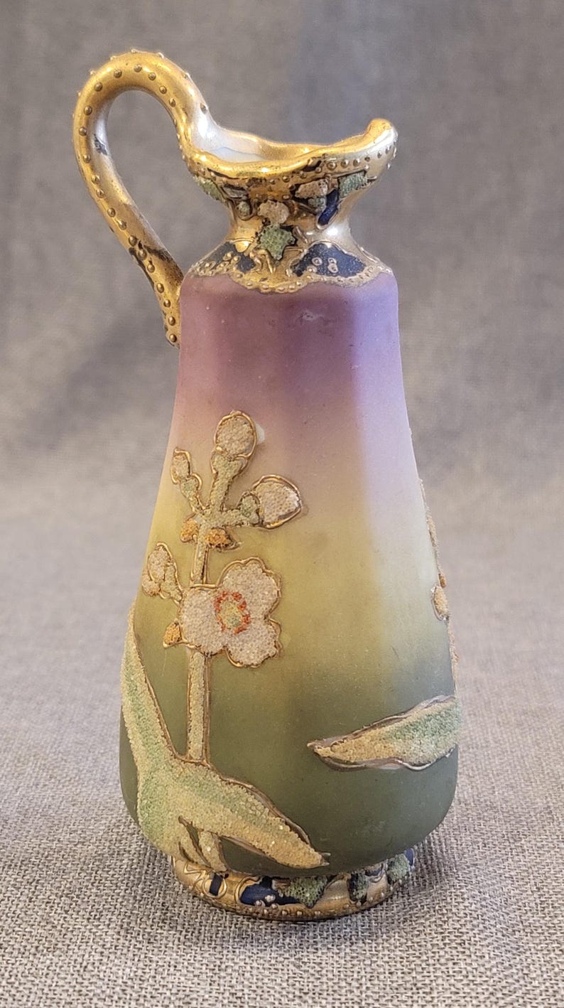 May include: A small, hand-painted porcelain pitcher with a gold handle and floral design. The pitcher has a gradient of purple, yellow, and green.