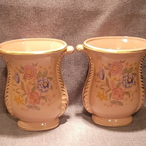 Puede incluir: Dos jarrones melocotón a juego con diseños florales. Cada jarrón tiene un borde y asas doradas, y presenta un patrón floral decorativo en rosa, azul y amarillo. Los jarrones son probablemente para fines decorativos.