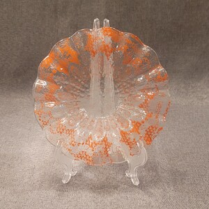 Op de afbeelding: Decoratieve heldere glazen plaat met een oranje gespikkeld ontwerp, tentoongesteld op een heldere plastic standaard. De plaat heeft een geschulpte rand en een getextureerd oppervlak. Het oranje patroon is geconcentreerd in het midden en langs de randen.