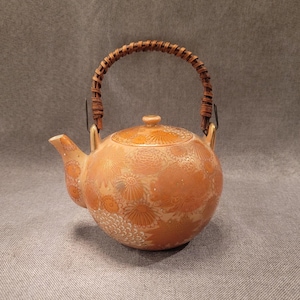 May include: A round, orange ceramic teapot with a floral design and a woven handle. The teapot has a spout and a lid with a small knob. The handle is made of woven brown material. The teapot is likely vintage or antique.