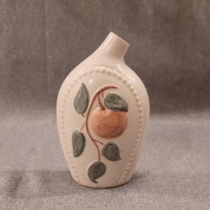 May include: Cream-colored ceramic vase with a decorative oval panel featuring a peach and green leaves. The vase has a curved shape with a narrow opening and a beaded border around the panel.