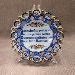 Könnte beinhalten: Ein dekorativer Keramikteller mit gewelltem Rand, verziert mit blau-weißen Blumenmustern und portugiesischem Text. Der Teller wird auf einem transparenten Kunststoffständer präsentiert.