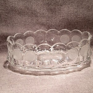 May include: Clear glass oval-shaped bowl with a scalloped rim. The bowl features etched coin-like designs around the sides and a sunburst pattern on the base. A decorative piece for serving or display.