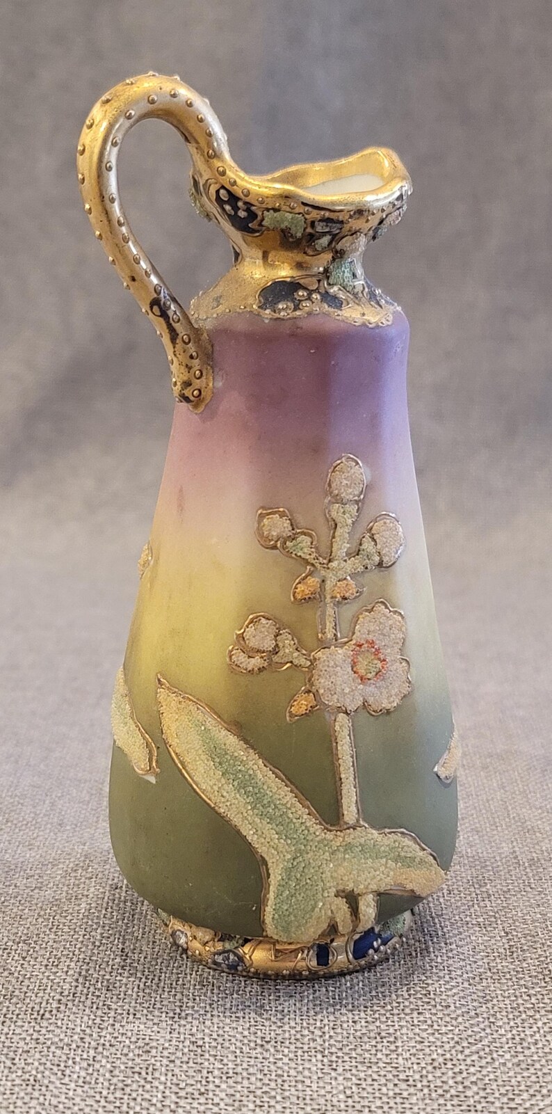 May include: A small, hand-painted porcelain pitcher with a gold handle and floral design. The pitcher is decorated with a green, yellow, and purple gradient and features a delicate floral motif with gold accents.
