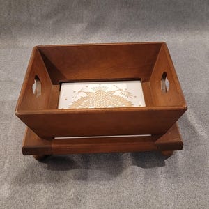 May include: A wooden serving tray with a square shape and a decorative ceramic tile insert. The tray has a raised base and cut-out handles. The tile features a gold-colored floral design on a white background. The wood has a warm, brown tone.