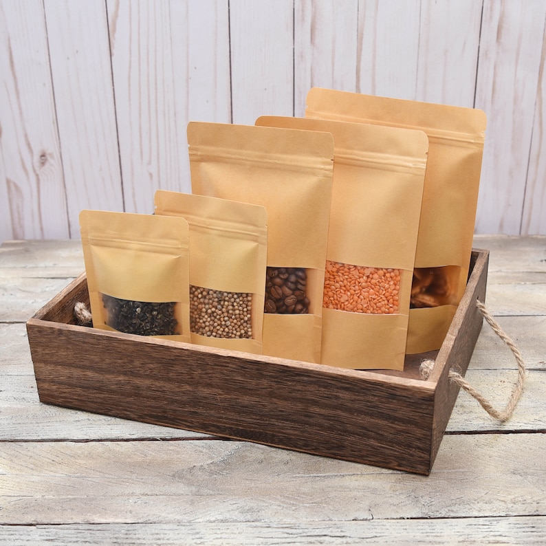 May include: Four brown paper bags with clear windows filled with different types of dried goods. The bags are sitting in a wooden box with rope handles.