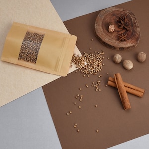 May include: A brown paper bag filled with coriander seeds, scattered on a brown surface with cinnamon sticks, whole nutmeg, and a wooden coaster with a star anise.