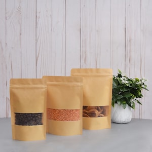 May include: Three brown paper bags with clear viewing windows. The bags are filled with coffee beans, lentils, and dried fruit. The bags are on a white wood surface.