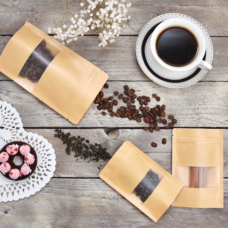 May include: Three brown paper pouches with clear windows, two filled with tea leaves and one with cinnamon sticks. The pouches are on a wooden surface with coffee beans scattered around them.