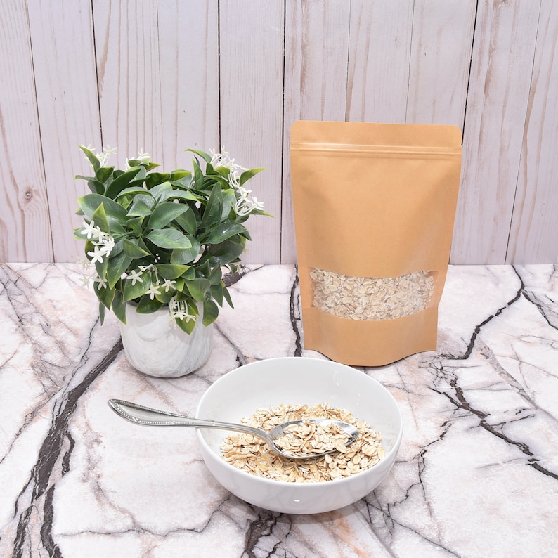 May include: A brown paper bag with a clear window showing rolled oats inside. A white bowl with rolled oats and a silver spoon is on a marble surface. A small potted plant with white flowers is in the background.