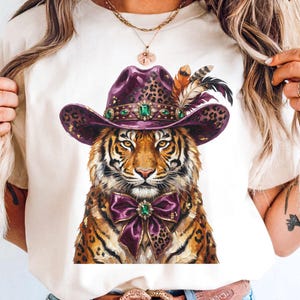 May include: A cream-colored t-shirt featuring a graphic print of a tiger wearing a purple leopard-print cowboy hat adorned with feathers and a green gemstone, and a matching purple leopard-print bow tie with a central green gemstone.