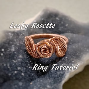 May include: A copper wire ring featuring a rose and leaf design. The ring is displayed on a grey background. Leafy Rosette Ring Tutorial.