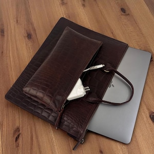 May include: A dark brown, textured leather laptop sleeve and matching accessory pouch. A silver laptop is partially visible inside the sleeve. A white charger is visible inside the accessory pouch. The items are on a wooden surface.