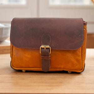 May include: A rectangular leather bag with a brown flap and a golden-yellow body. The bag features a brown strap with a gold-colored buckle. The bag is sitting on a wooden surface.