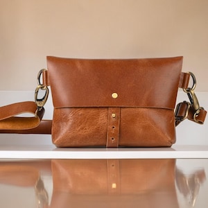 May include: A brown leather crossbody bag with a flap closure and adjustable strap. The bag features brass-colored hardware and a button closure. The bag is sitting on a white surface.