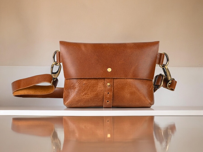 May include: A brown leather crossbody bag with a flap closure and adjustable strap. The bag features brass-colored hardware and a button closure. The bag is sitting on a white surface.