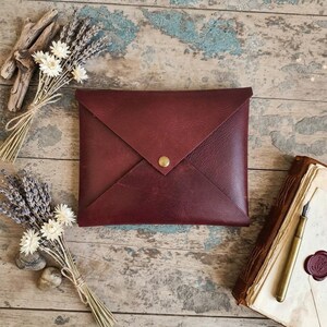 Op de afbeelding: Een bordeauxrode leren envelop-stijl clutch met een gouden drukknoopsluiting. De clutch wordt omringd door gedroogde lavendel en een vintage dagboek met een pen. De items zijn gerangschikt op een rustiek, verweerd houten oppervlak.