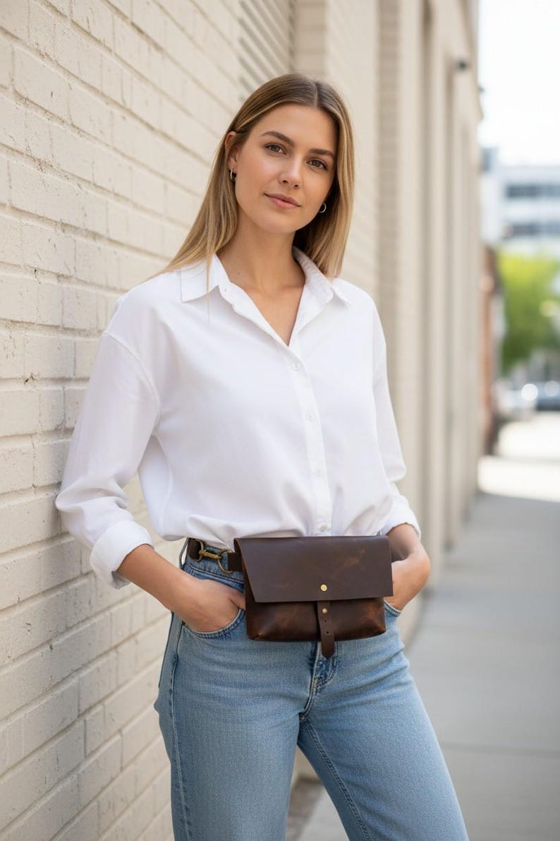 May include: A brown leather fanny pack with a flap closure and a brass button. The bag is worn around the waist with a belt. The person is wearing a white button-down shirt and blue jeans.