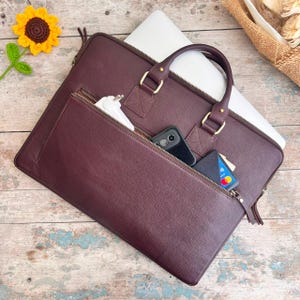 Può includere: Una borsa per computer portatile in pelle color bordeaux scuro con una grande tasca frontale con cerniera, due manici superiori e finiture dorate. Lo scomparto principale contiene un computer portatile argento e una carta di credito.