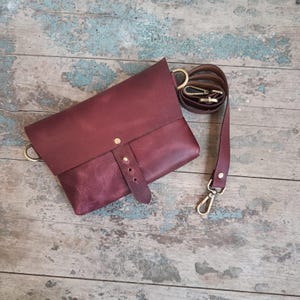 May include: A burgundy leather crossbody bag with a flap closure and adjustable strap. The bag features gold-colored hardware, including a clasp and D-rings. The strap is made of matching burgundy leather.