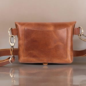 May include: A brown leather fanny pack with a front pocket and adjustable strap. The bag features brass-colored hardware, including a clasp and D-rings. The leather has a natural, textured appearance.