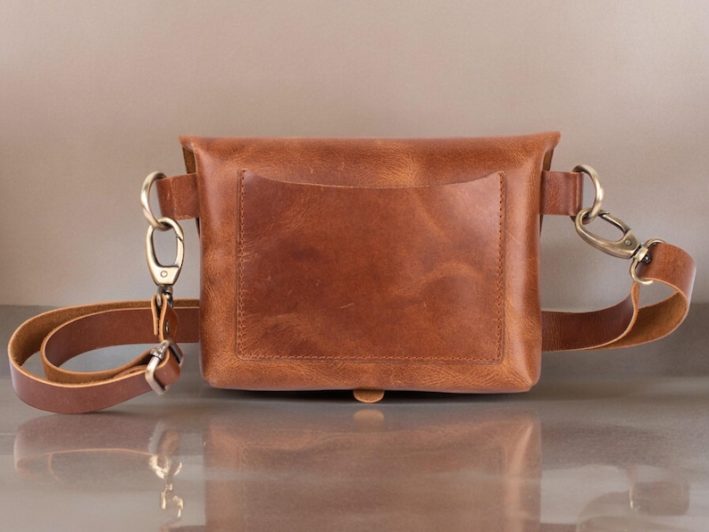 May include: A brown leather fanny pack with a front pocket and adjustable strap. The bag features brass-colored hardware, including a clasp and D-rings. The leather has a natural, textured appearance.