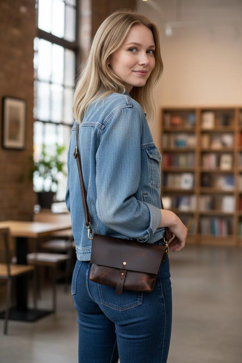 May include: A dark brown leather crossbody bag with a flap closure and adjustable strap. The bag features a small front pocket and gold-colored hardware. The person in the image is wearing a denim jacket and blue jeans.