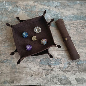 Peut inclure: Un plateau à dés carré en cuir marron foncé avec des coins à boutons-pression et un étui à dés cylindrique assorti en cuir. À l'intérieur du plateau se trouvent quatre dés de jeu à facettes bleus, violets, transparents et dorés.