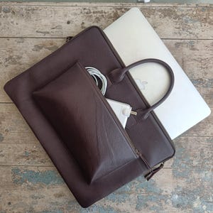 May include: A dark brown leather laptop bag with a matching smaller pouch. A silver laptop is partially visible, along with a white charger and cable. The bag has a handle and a zipper closure. The pouch is zippered and contains a white charger and cable.