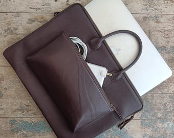 May include: A dark brown leather laptop bag with a matching smaller pouch. A silver laptop is partially visible, along with a white charger and cable. The bag has a handle and a zipper closure. The pouch is zippered and contains a white charger and cable.