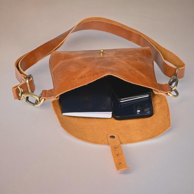 May include: A brown leather crossbody bag with an adjustable strap and a flap closure. The bag is open, revealing a black smartphone and a small notebook inside. The bag has brass-colored hardware.