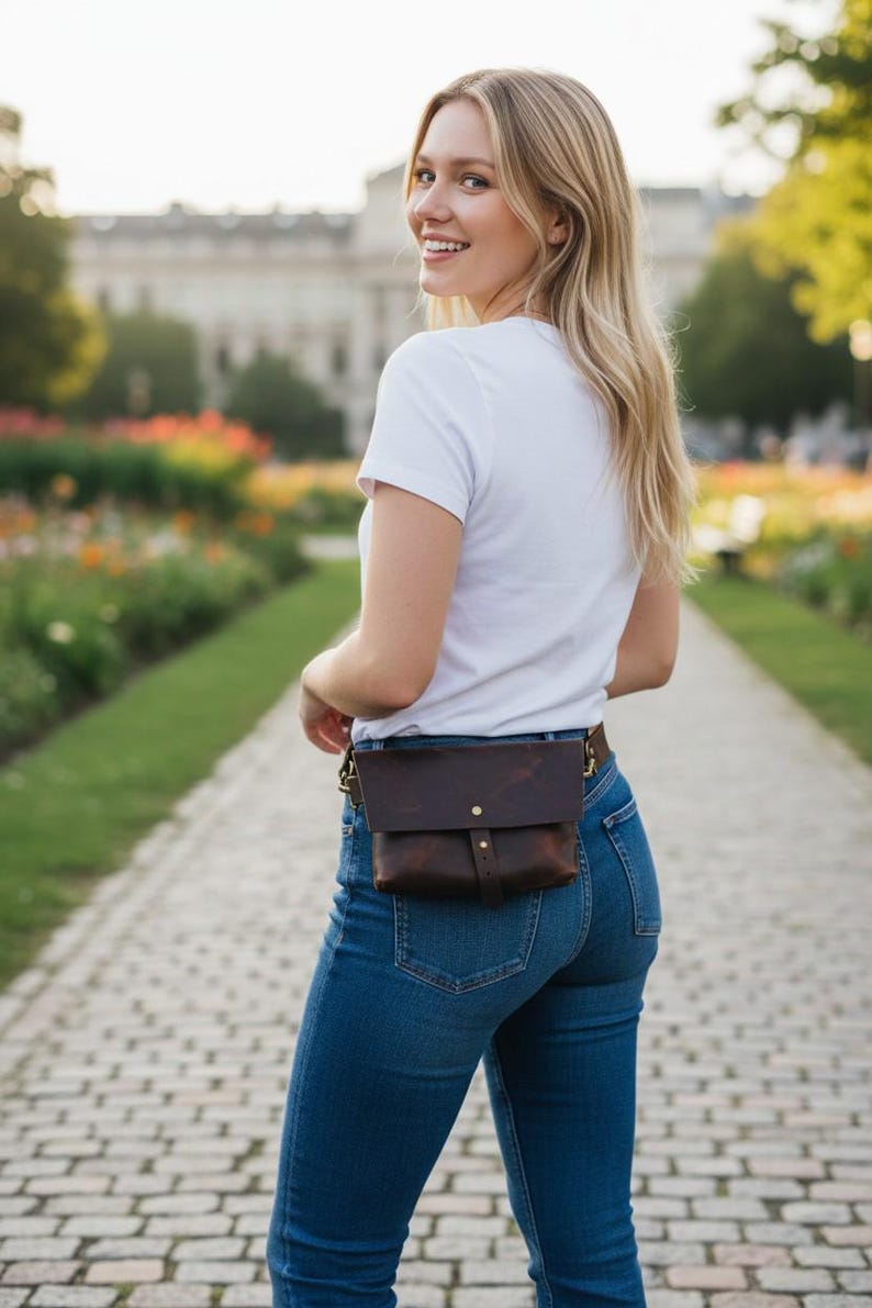 May include: A brown leather fanny pack with a flap closure and brass hardware. The bag is worn around the waist with a belt. The person is wearing a white t-shirt and blue jeans. The background is a park with a stone path.
