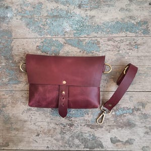 May include: A burgundy leather crossbody bag with a flap closure and adjustable strap. The bag features brass-colored hardware, including D-rings and a clasp. The strap is detachable. The bag is shown on a weathered wooden surface.