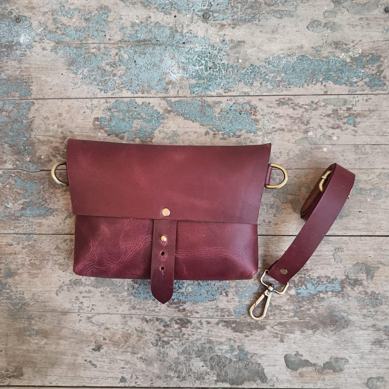 May include: A burgundy leather crossbody bag with a flap closure and adjustable strap. The bag features brass-colored hardware, including D-rings and a clasp. The strap is detachable. The bag is shown on a weathered wooden surface.