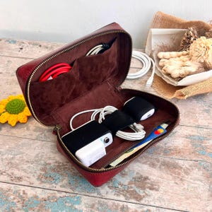 May include: A deep red leather zipped organiser case with a dark brown suede interior. The case contains multiple compartments holding white and red charging cables, a white power adapter, a black wireless earbud case, and a payment card.
