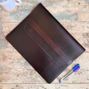 May include: A dark brown leather portfolio with a darker brown strap closure. The portfolio is rectangular with rounded corners. Two pens with blue and purple ink are next to the portfolio.