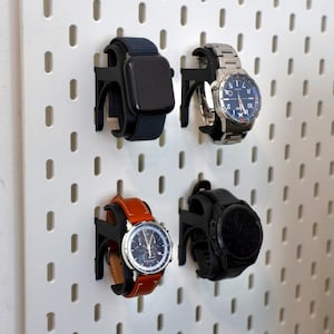 May include: Four black plastic watch holders mounted on a white pegboard. Each holder supports a different timepiece: a smartwatch with a dark band, a silver-tone analog watch, a watch with a tan leather strap, and a black digital sports watch.