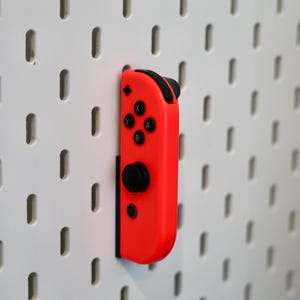 May include: A bright neon red video game controller component featuring a black analog stick, a directional pad, and four circular buttons labeled with the letters A, B, X, and Y. The controller is mounted vertically against a white pegboard wall.
