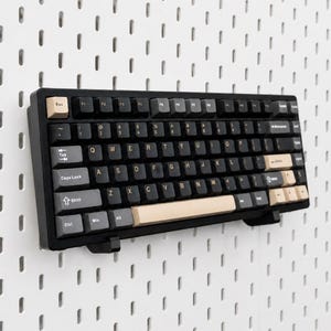 May include: A black mechanical keyboard featuring dark gray keycaps with white lettering, accented by light beige keys for the Esc, spacebar, and right-side function keys. The keyboard is mounted horizontally on a white pegboard wall panel.