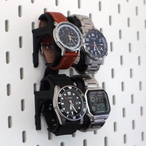 Puede incluir: Cuatro relojes de pulsera expuestos en una pared perforada blanca. Los relojes tienen varios materiales de correa, incluyendo cuero y metal. Las esferas de los relojes son negras y plateadas, con diferentes estilos de manecillas y números. Los relojes se sujetan con soportes negros.