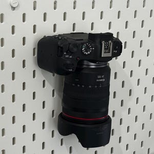 May include: A black Canon camera with a 24-105 lens is mounted on a white pegboard. The lens has a black lens hood. The camera's controls are visible on top. The pegboard has evenly spaced rectangular holes.